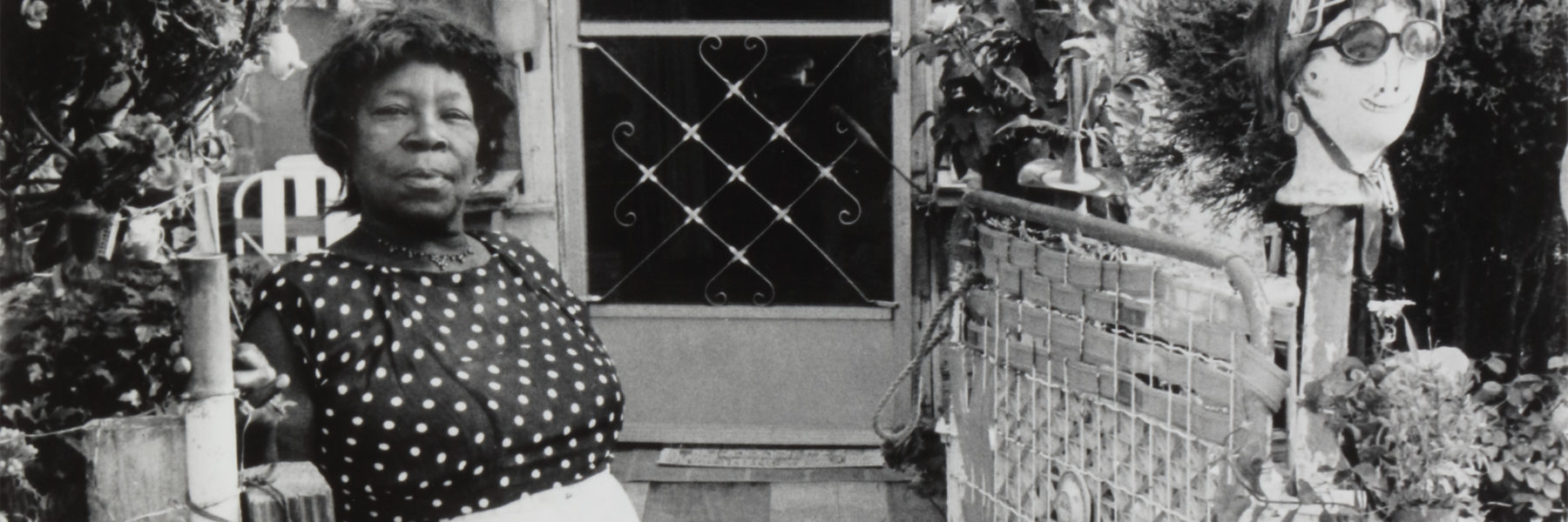 Nellie Mae Rowe wears a black shirt with white polkadots and stands in front of her home to the left of her front door. To the right of the door is a doll head wearing sunglasses perched on a post.