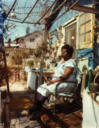 Nellie Mae Rowe sits in a chair on the porch in front of her house under an arbor. The yard in the background contains several small trees covered with multi-colored ornaments.