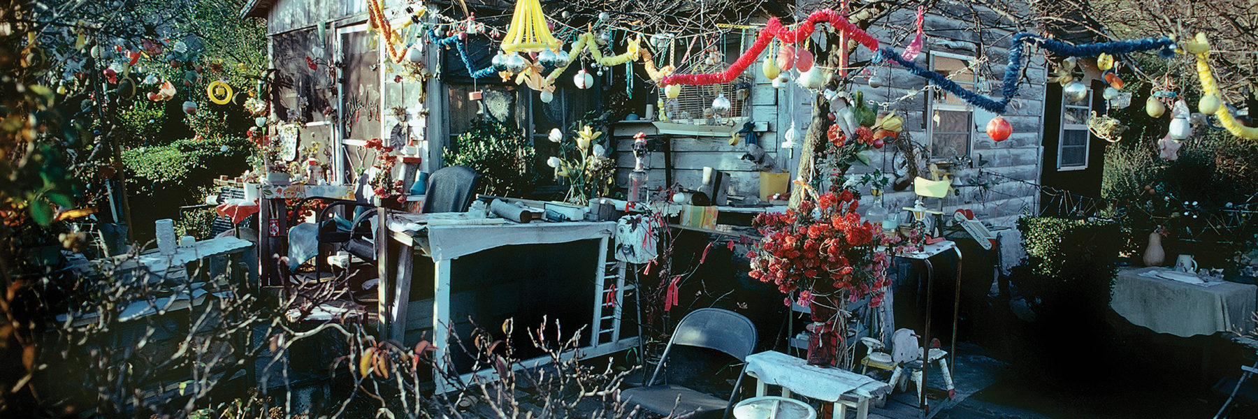 A house stands at the center of the photograph, a bare tree in the courtyard in front of the house. The tree branches are decorated with a myriad of colorful ornaments and baubles. The courtyard is full of folding chairs and tables covered with an assortment of projects and flower arrangements.