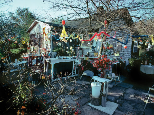 A house stands at the center of the photograph, a bare tree in the courtyard in front of the house. The tree branches are decorated with a myriad of colorful ornaments and baubles. The courtyard is full of folding chairs and tables covered with an assortment of projects and flower arrangements.