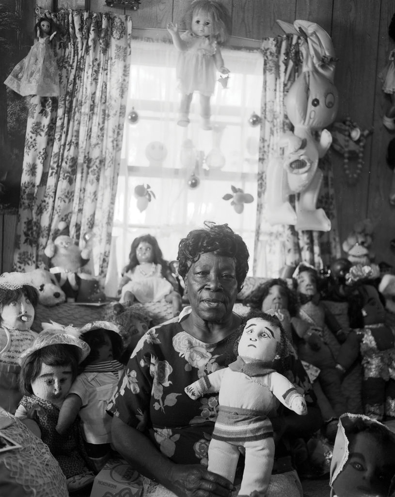 Nellie Mae Rowe wearing a flowered dress sits on a sofa holding a stuffed doll in her lap. On the seat and back of the sofa behind her are numerous dolls and stuffed animals. Two dolls and a blow-up rabbit hang from the curtain rod of the window behind her.