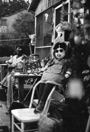 Nellie Mae Rowe sits in a chair on her porch in the background of a black and white photograph. In the foreground, a stuffed doll wearing a gingham dress, sunglasses, and dark tights with shoes sits on the top of a white wooden chair. The small table between them is covered with sea shells, an ornamental clock, and a toy horse.