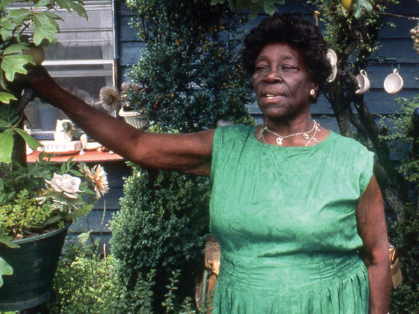Nellie Mae Rowe wears a green dress and stands in the garden in front of her house. Her right hand rests on the branch of a tree. The tree branches above her head are decorated with fruit-shaped ornaments, and a rack holding teacups hangs from the tree to the right of Rowe's head.