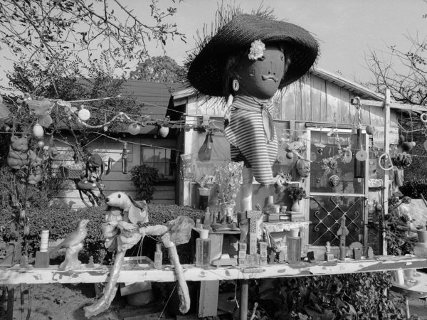 A black and white photograph of the front of Nellie Mae Rowe's house depicts the variety of objects decorating her front yard. A large doll's head wearing a straw hat and a striped dickie stands atop an ornament-covered wire in front of the house. Below it, a wooden rail is covered with a collection of objects including a toy car, a ceramic bird, building blocks, and a dog made of pieces of plastic. Many more objects hang from tree branches and wires in front of the house.