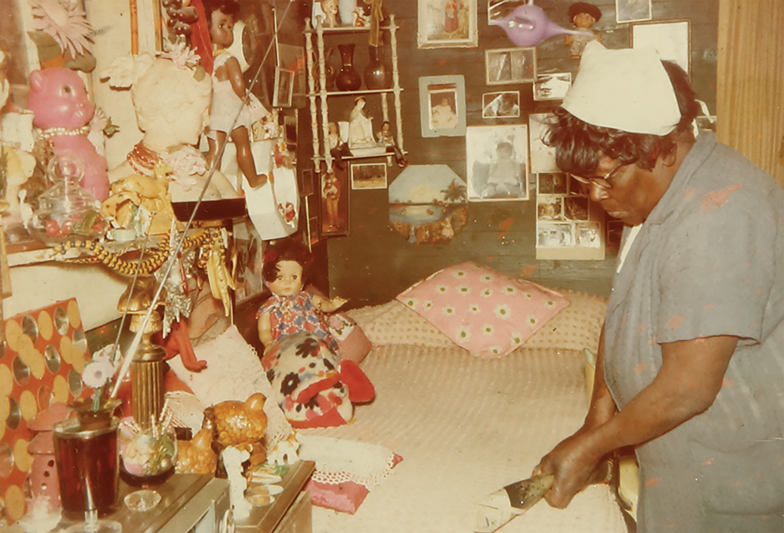 Photograph of Nellie Mae Rowe in a blue dress and white kerchief on her hair standing next to a bed and rolling some paper in her hands. The walls around the bed are heavily decorated with photographs, dolls, beads, and other collectible items.