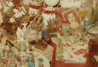 Photograph of a dresser half covered with decorations. A chewing gum sculpture of a crouching animal stands at the front of the dresser, with another sculpture of a figure covered in multi-colored balls. A drawn portrait of a woman against a red background is placed to the right behind a stuffed animal and boot-shaped toy on wheels.