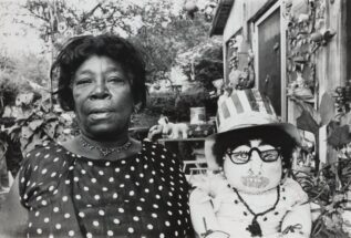 Nellie Mae Rowe stands outside her home in a polka dot blouse beside a doll wearing a striped top hat that says “PEACE”, vintage librarian glasses, and a corsage.